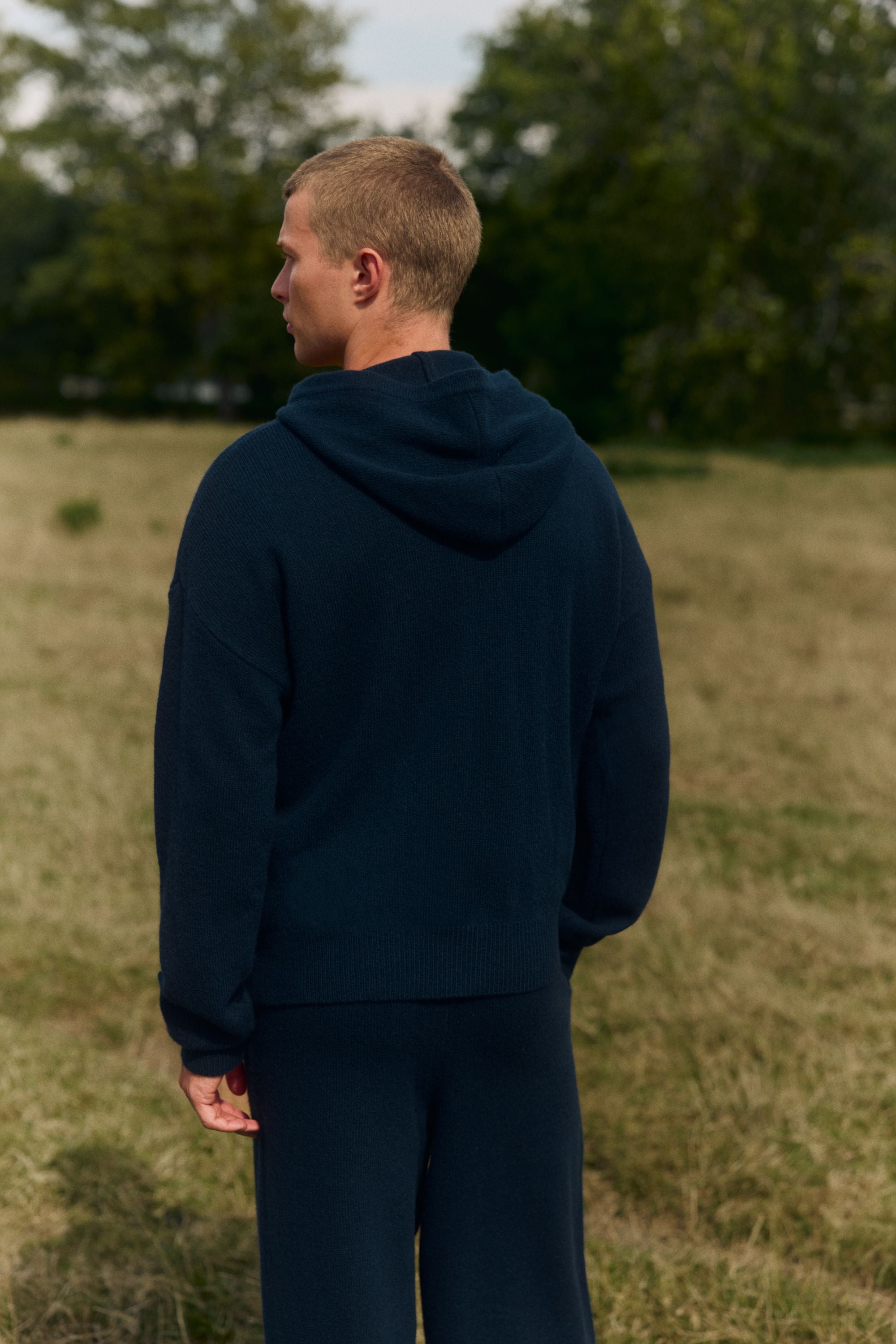 Man viewed from behind wearing a navy CASPARA woolknit hooded jacket in a grassy field.